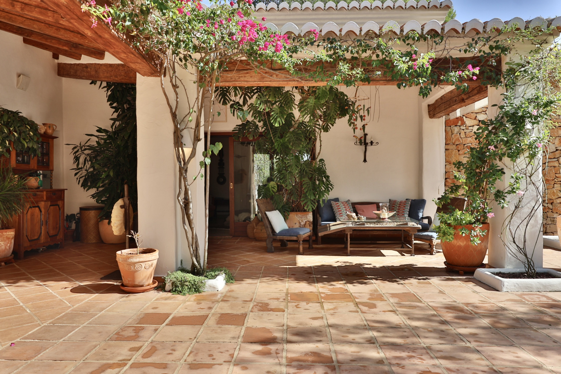 Terraza cubierta con suelo de baldosas de terracota, vigas de madera expuestas y una pérgola adornada con buganvillas. Incluye zona de estar exterior y pared de piedra rústica. Terraza cubierta con suelo de baldosas de terracota, vigas de madera expuestas y una pérgola adornada con buganvillas. Incluye zona de estar exterior y pared de piedra rústica.