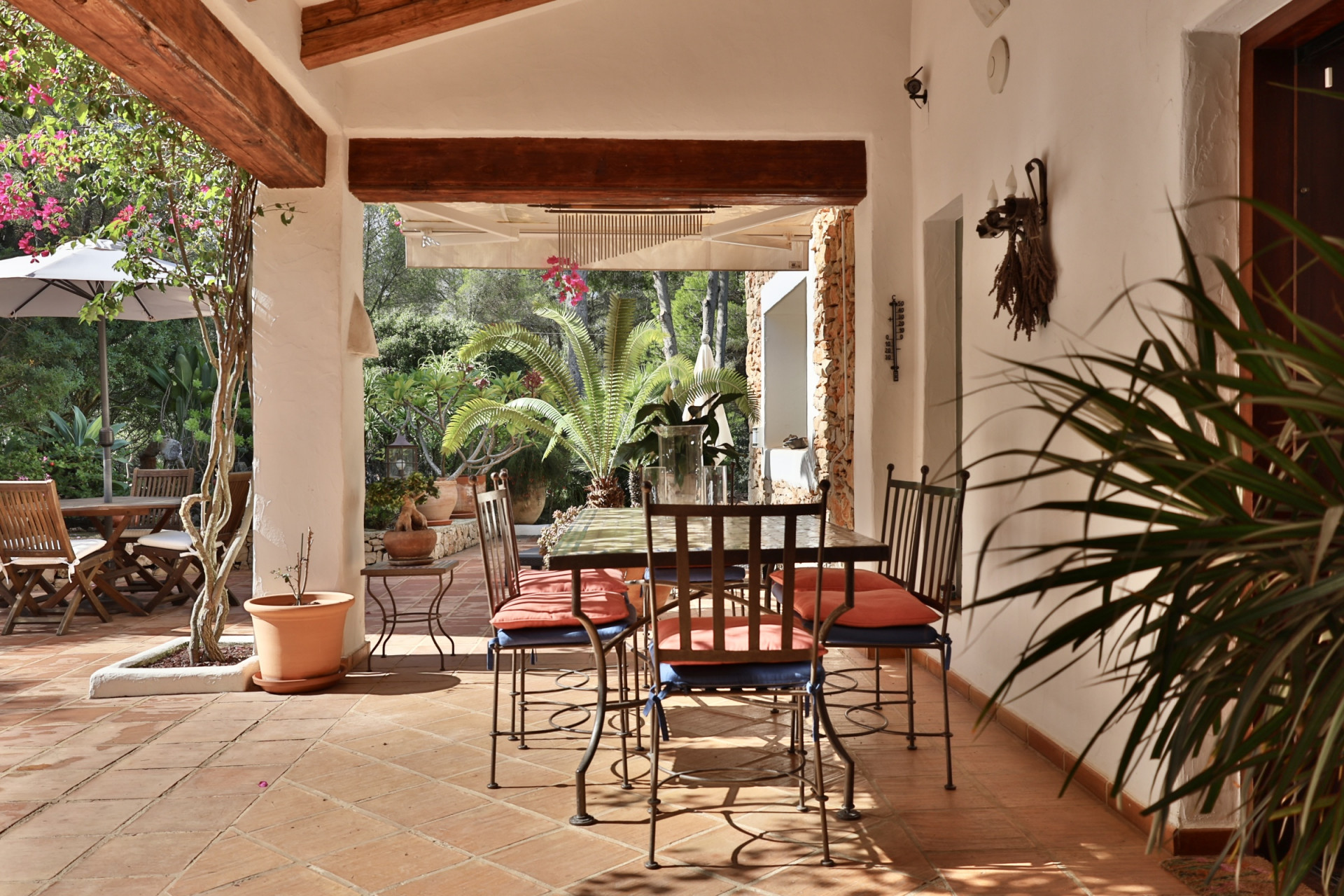 Terraza cubierta con suelo de terracota y vigas de madera, equipada con mesa de comedor de hierro forjado y vistas al exuberante jardín mediterráneo. Terraza cubierta con suelo de terracota y vigas de madera, equipada con mesa de comedor de hierro forjado y vistas al exuberante jardín mediterráneo.