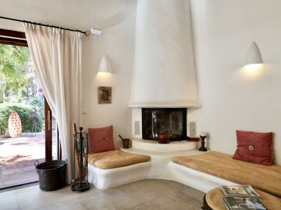 Acogedora sala de estar con chimenea de estuco blanco y bancos integrados, ofreciendo acceso directo a un patio exterior con jardín y luz natural.