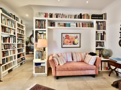 Acogedora sala de lectura con sofá rosa, estanterías empotradas repletas de libros y un cuadro floral. Suelo de baldosas claras y lámpara de mesa.