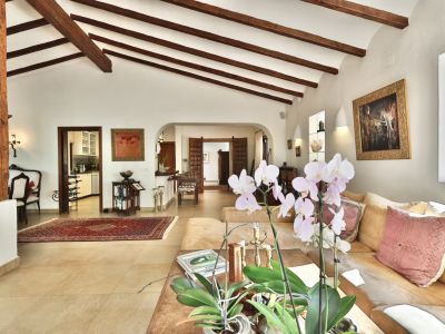 Amplio salón con techo abovedado y vigas de madera a la vista, columna rústica, suelo de gres claro y sofá seccional. Conexión a la cocina y otras áreas a través de arcos.