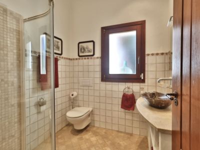 Baño con ducha de mosaico, inodoro suspendido y lavabo de piedra con grifería cromada. Ventana de madera y azulejos blancos con cenefa decorativa.