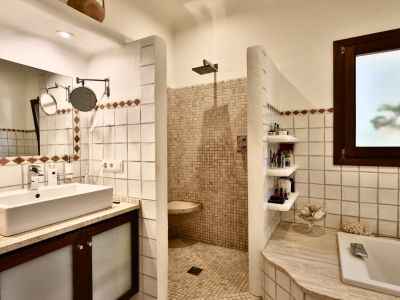 Baño con lavabo rectangular sobre mueble de madera oscura y encimera de piedra clara. Ducha de obra con mosaico beige y bañera integrada. Ventana esmerilada.