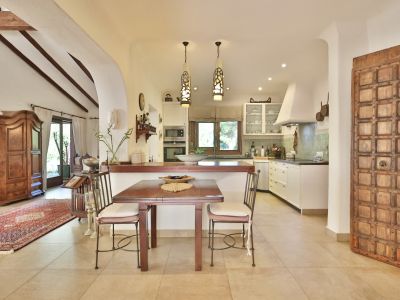 Cocina abierta con encimeras oscuras, azulejos verdes y electrodomésticos integrados. Mesa de comedor de madera junto a una puerta decorativa y un salón con vigas vistas.