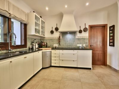 Cocina equipada con armarios blancos de estilo tradicional, encimeras oscuras y azulejos verdes. Destaca la ventana de madera con persiana romana y la puerta de madera maciza.