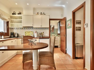 Cocina funcional con encimeras de madera, armarios blancos y suelo de terracota. Incluye una mesa redonda con sillas de mimbre y acceso a un baño con azulejos verdes.