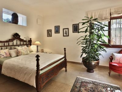 Dormitorio con cama de madera tallada y cabecero ornamentado. Incluye un sillón de terciopelo rojo, una planta grande en maceta y una ventana con rejas que ofrece vistas al exterior.