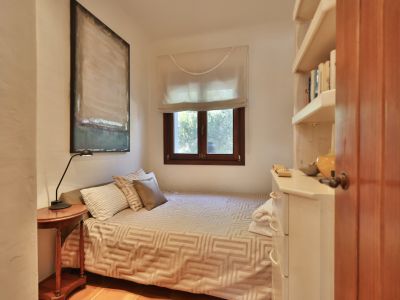 Dormitorio con cama individual, mesita de noche de madera y lámpara. Ventana con marco de madera y persiana, estantería blanca con libros y cómoda. Cuadro abstracto y suelo de terracota.