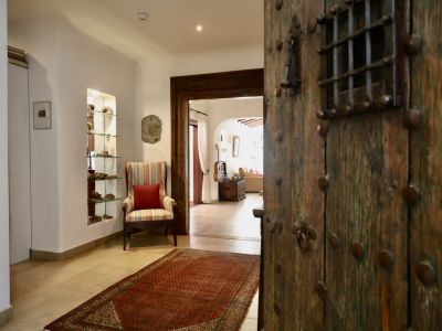 La rústica puerta de entrada de madera con herrajes metálicos y reja se abre a un luminoso recibidor. Destacan un sillón a rayas, una hornacina con estantes de cristal y una alfombra roja sobre suelo de baldosas.