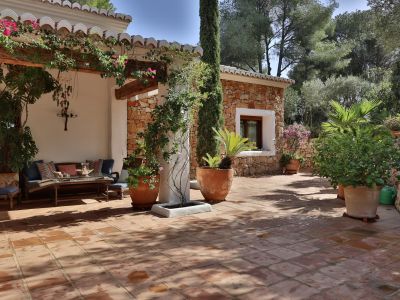 Patio con baldosas de terracota, pérgola sombreada con buganvillas, zona de asientos exterior y anexo de piedra entre plantas mediterráneas y ciprés.