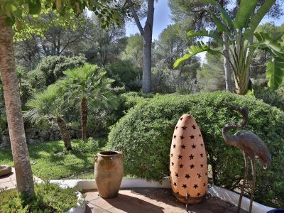 Patio de jardín con suelo de terracota, destacando una maceta rústica, una lámpara de estrellas y una estatua de garza, enmarcado por exuberante vegetación mediterránea y palmeras.