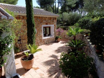 Patio exterior con baldosas de terracota junto a una edificación de fachada de piedra. Incluye un ciprés, plantas mediterráneas en macetas y un muro de piedra que delimita la vegetación.