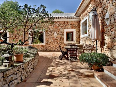 Patio exterior con suelos de terracota y muros de piedra vista, equipado con mobiliario de madera y sombrilla. Vegetación en macetas y un árbol proporcionan sombra natural.