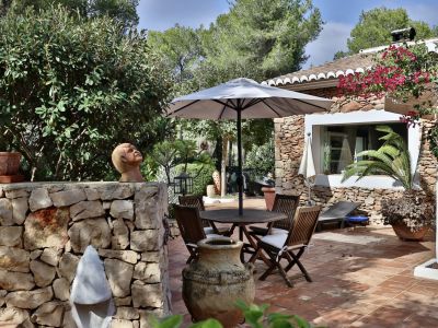 Patio exterior de terracota con mesa de madera y sombrilla, rodeado de un muro de piedra y exuberante vegetación mediterránea, junto a la fachada de piedra.