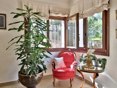 Rincón acogedor con ventanales de madera que ofrecen vistas al jardín, sillón rojo tapizado y una planta exuberante. Incluye mesa auxiliar con estatua de Buda y orquídea.