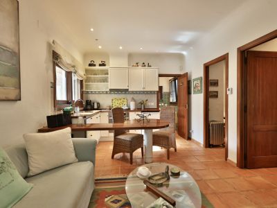 Salón-cocina de concepto abierto con suelo de terracota. La cocina equipada presenta muebles blancos, encimera de madera y barra de desayuno integrada con sillas de mimbre.
