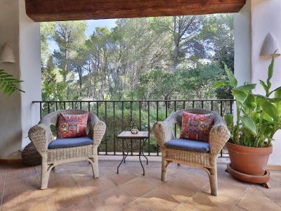 Terraza cubierta con dos sillones de mimbre y cojines azules, mesa auxiliar de forja, suelo de terracota y vistas a la exuberante vegetación natural.