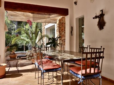Terraza cubierta con mobiliario de comedor de hierro forjado y mesa con patrón, sobre suelo de baldosas de terracota. Ofrece vistas a un exuberante jardín mediterráneo y muro de piedra.