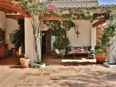 Terraza cubierta con suelo de baldosas de terracota, vigas de madera expuestas y una pérgola adornada con buganvillas. Incluye zona de estar exterior y pared de piedra rústica.