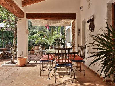 Terraza cubierta con suelo de terracota y vigas de madera, equipada con mesa de comedor de hierro forjado y vistas al exuberante jardín mediterráneo.