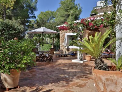 Terraza exterior con suelo de terracota, mobiliario de comedor de madera bajo sombrilla y exuberante vegetación mediterránea, destacando la bougainvillea en la fachada de piedra.