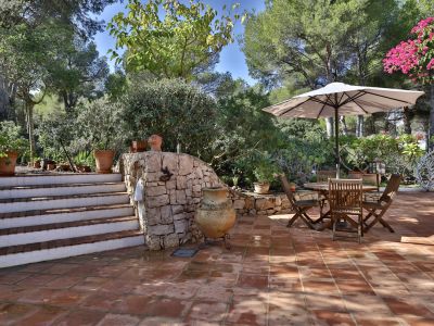 Terraza exterior con suelo de terracota, muro de piedra y escalera. Conjunto de mesa y sillas de madera bajo sombrilla, rodeado de vegetación mediterránea y una gran tinaja decorativa.