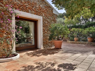 Terraza exterior con suelo de terracota y fachada de piedra seca, enmarcando una puerta acristalada de madera. Una bougainvillea en flor y plantas mediterráneas adornan el jardín soleado.