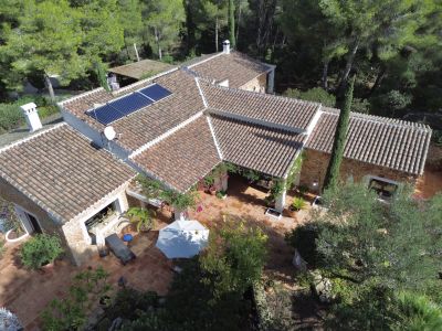 Vista aérea de la villa con tejados de teja, muros de piedra y una extensa terraza de terracota. Se aprecian paneles solares y una pérgola cubierta, rodeada de vegetación mediterránea.