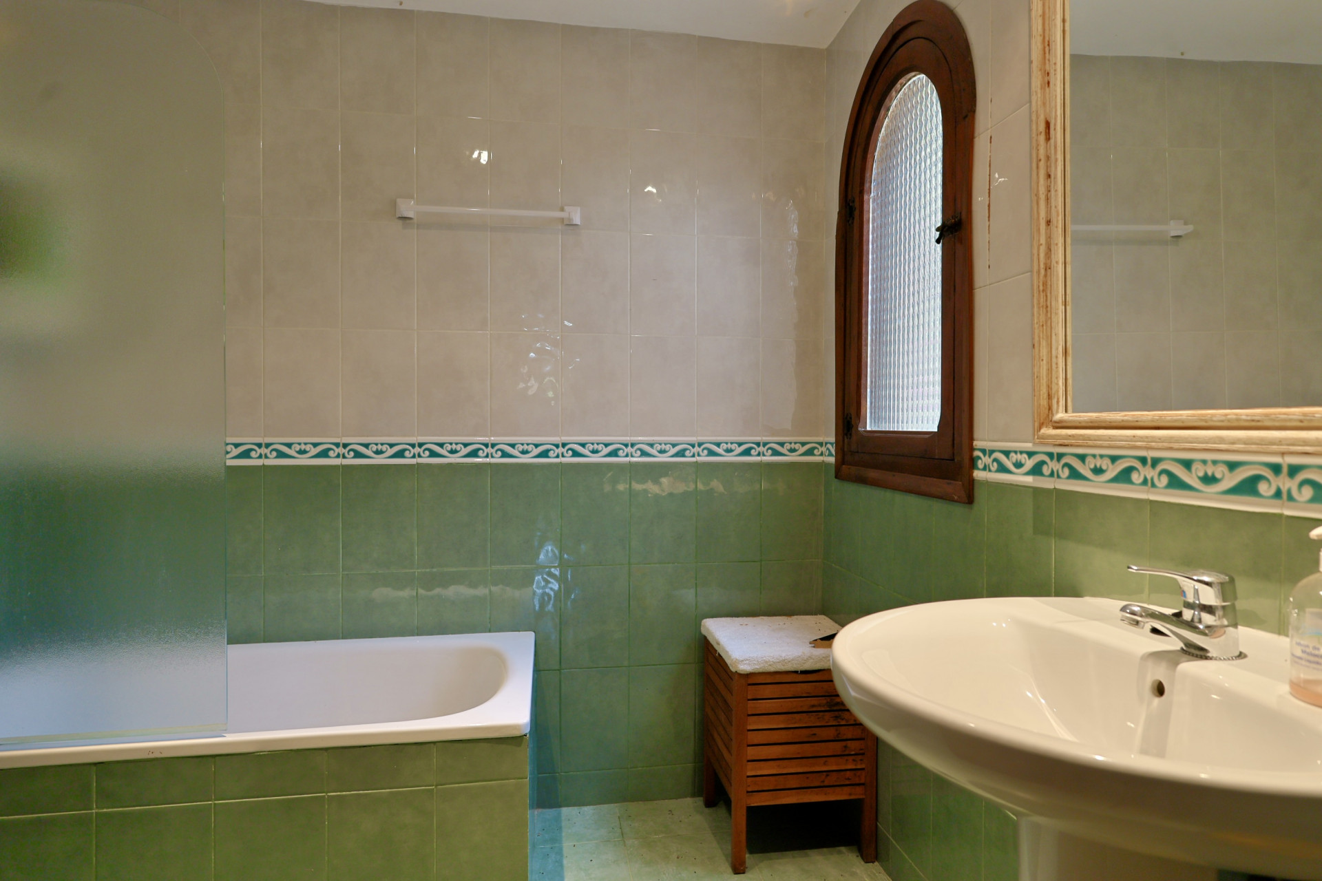 Baño con paredes alicatadas en tonos verde y beige, bañera con mampara de cristal esmerilado y lavabo de pedestal. Ventana de madera arqueada con cristal texturizado. Baño con paredes alicatadas en tonos verde y beige, bañera con mampara de cristal esmerilado y lavabo de pedestal. Ventana de madera arqueada con cristal texturizado.
