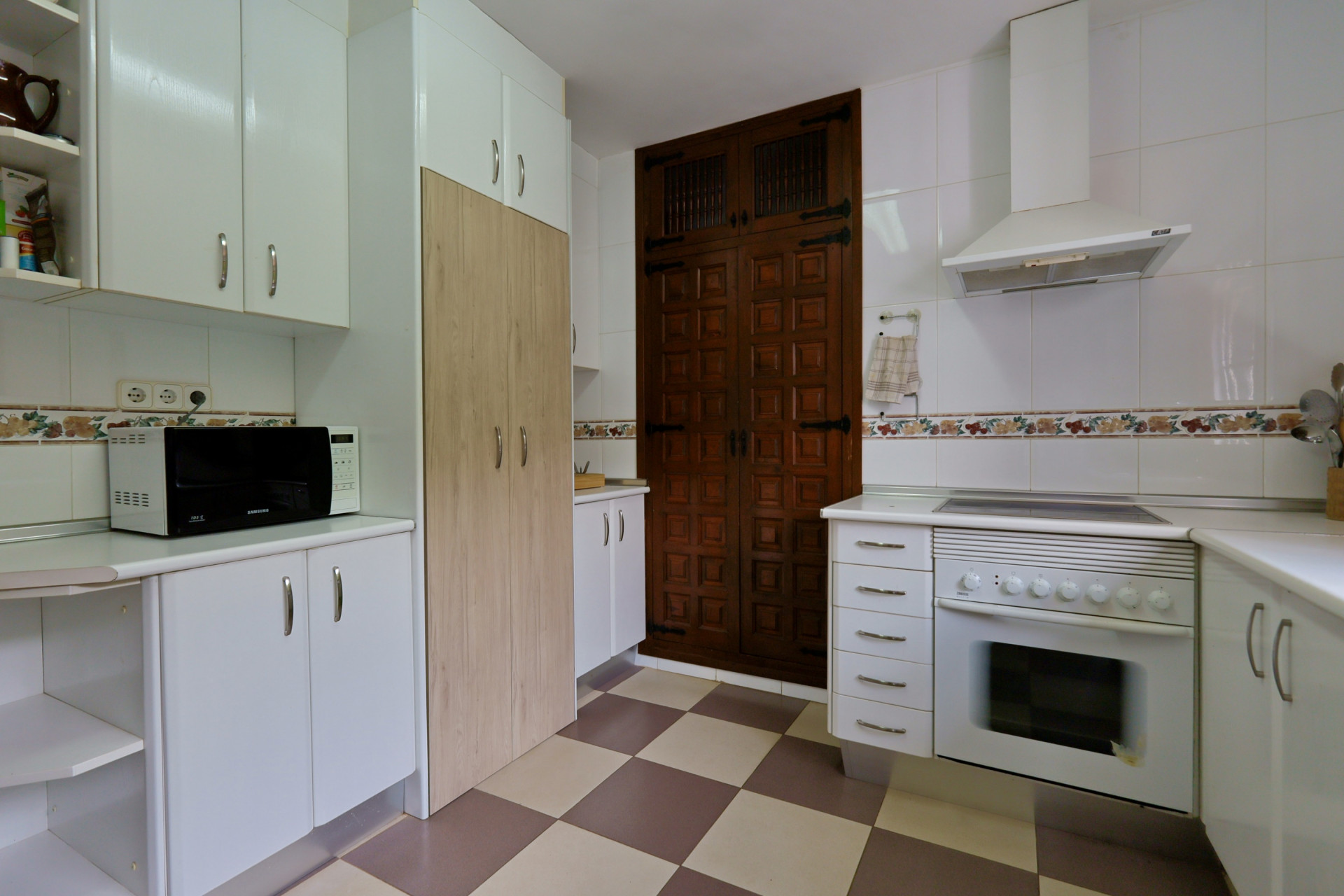 Cocina equipada con armarios blancos, microondas y horno integrado. Destaca la puerta de madera oscura con paneles y el suelo de baldosas ajedrezadas. Cocina equipada con armarios blancos, microondas y horno integrado. Destaca la puerta de madera oscura con paneles y el suelo de baldosas ajedrezadas.