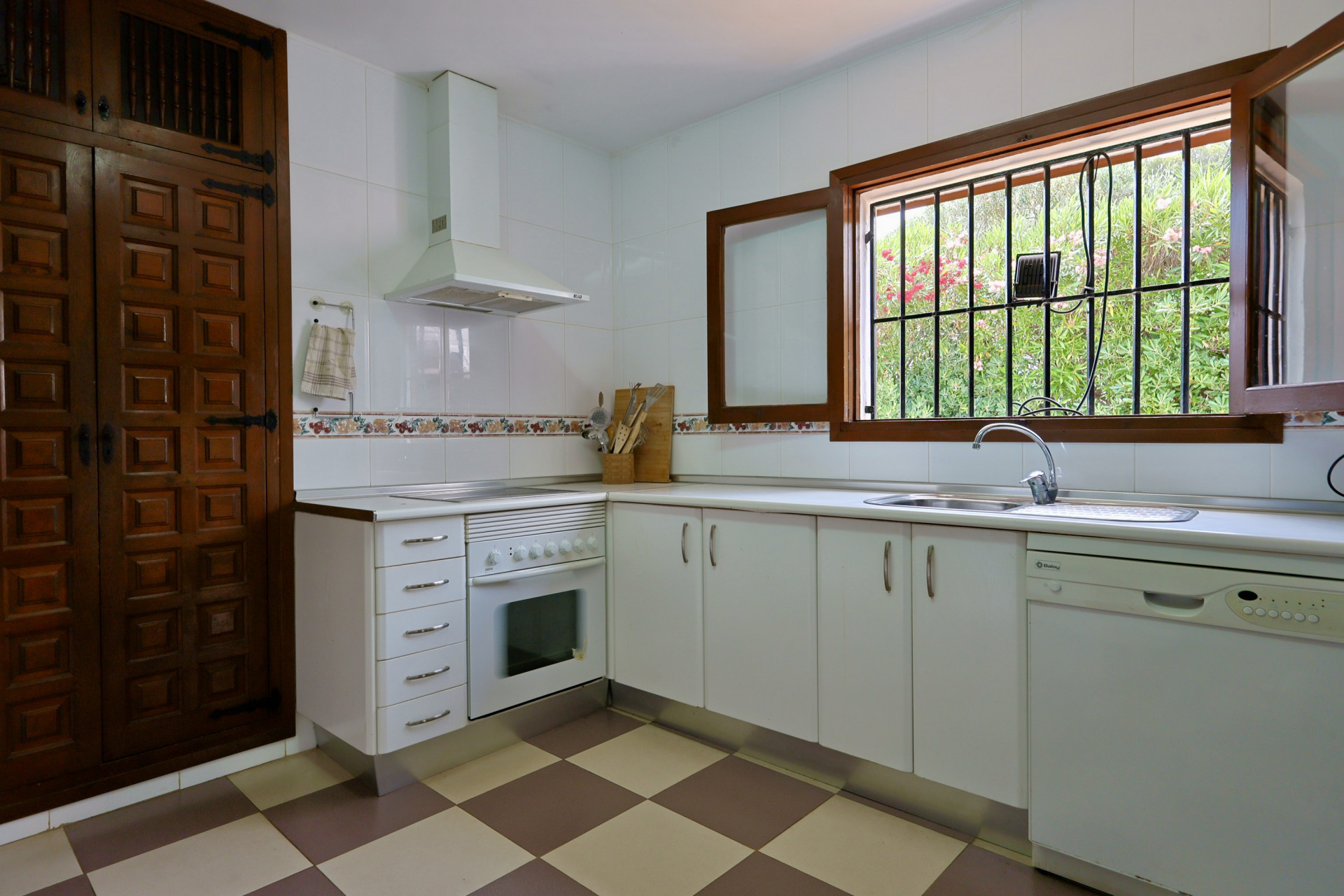 Cocina funcional con armarios blancos, suelo de baldosas a cuadros y una ventana de madera con rejas que ofrece vistas al jardín. Incluye fregadero de acero y lavavajillas. Cocina funcional con armarios blancos, suelo de baldosas a cuadros y una ventana de madera con rejas que ofrece vistas al jardín. Incluye fregadero de acero y lavavajillas.