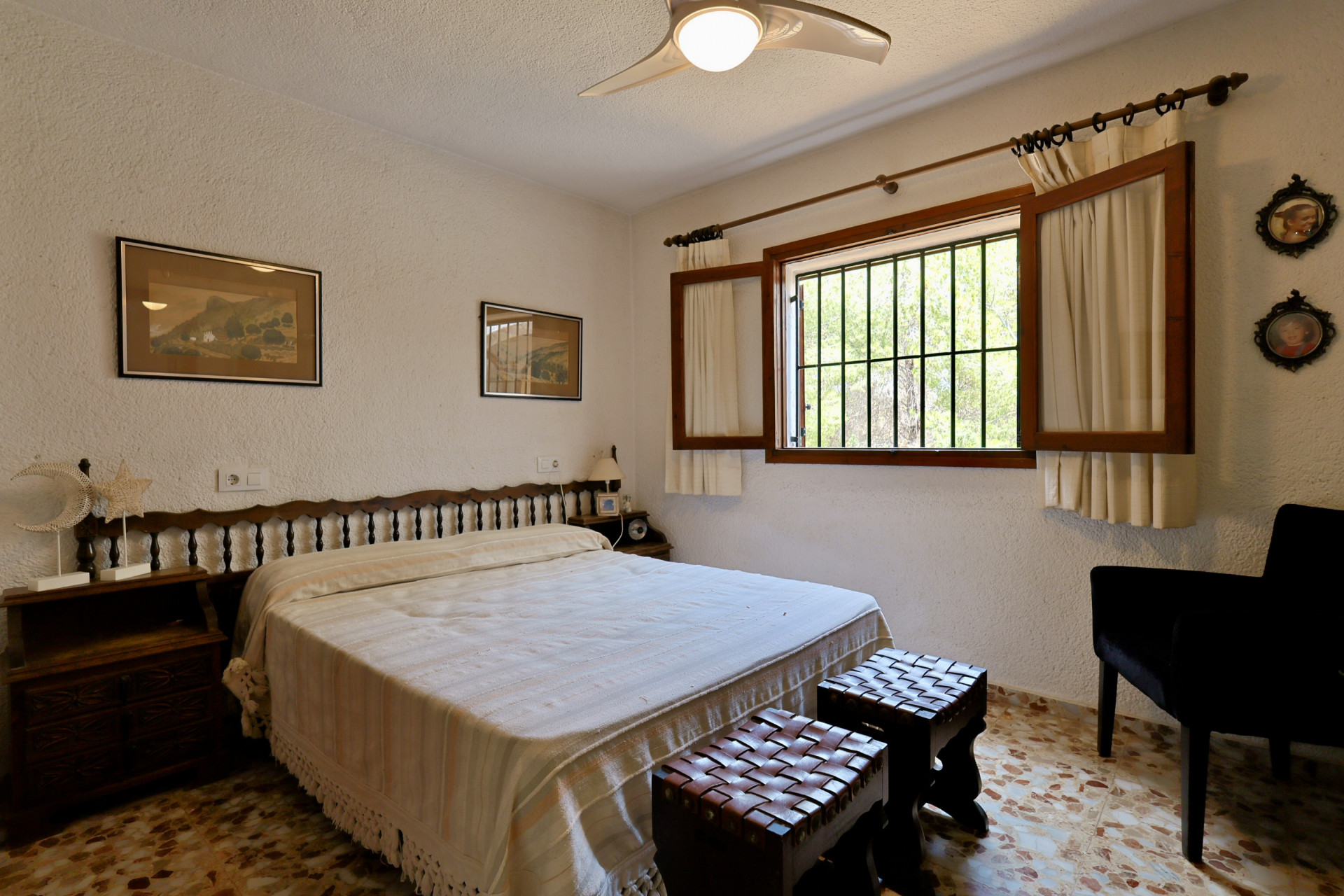 Dormitorio con cama doble, cabecero de madera y suelo de baldosas. La ventana con contraventanas y rejas ofrece luz natural y vista a la vegetación exterior. Dormitorio con cama doble, cabecero de madera y suelo de baldosas. La ventana con contraventanas y rejas ofrece luz natural y vista a la vegetación exterior.