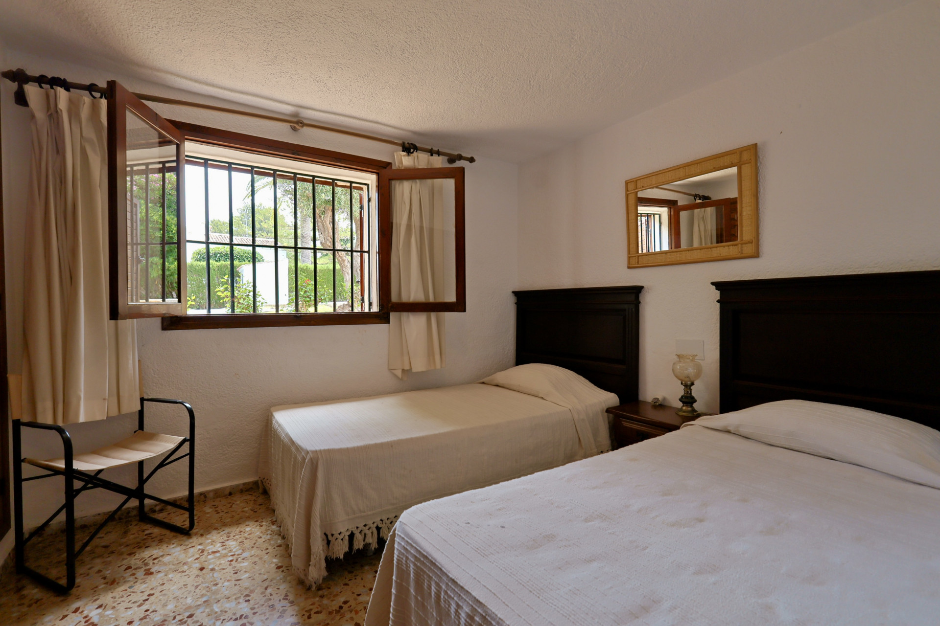 Dormitorio con dos camas individuales de cabeceros de madera oscura, mesita de noche compartida y ventana con rejas hacia el jardín. Suelo de terrazo y espejo con marco trenzado. Dormitorio con dos camas individuales de cabeceros de madera oscura, mesita de noche compartida y ventana con rejas hacia el jardín. Suelo de terrazo y espejo con marco trenzado.
