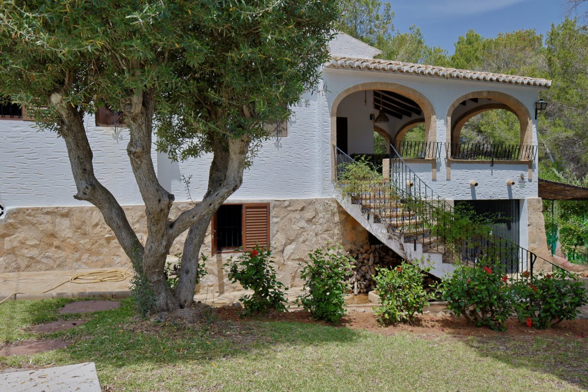 El exterior de la villa muestra una fachada blanca texturizada sobre una base de piedra, con una terraza cubierta de arcos y una escalera exterior de azulejos, junto a un olivo. El exterior de la villa muestra una fachada blanca texturizada sobre una base de piedra, con una terraza cubierta de arcos y una escalera exterior de azulejos, junto a un olivo.