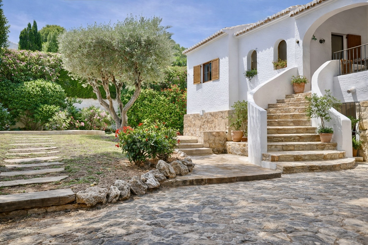 Fachada blanca de la villa con escalera curva de piedra que conduce a la entrada. El jardín mediterráneo presenta un olivo central, arbustos y flores, con un camino de losas. Fachada blanca de la villa con escalera curva de piedra que conduce a la entrada. El jardín mediterráneo presenta un olivo central, arbustos y flores, con un camino de losas.
