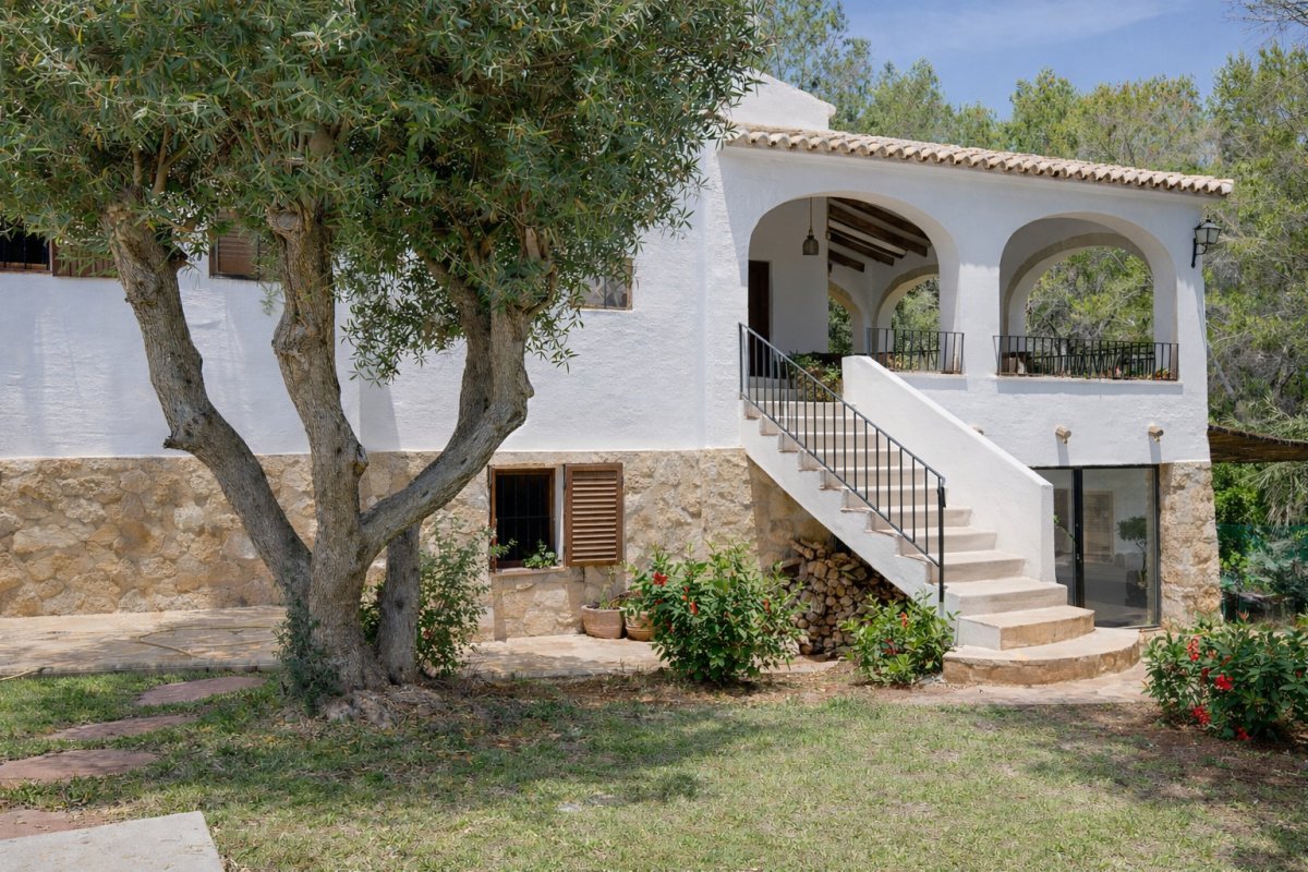 Fachada de villa con base de piedra y muros blancos, destacando un olivo maduro y una escalera exterior que conduce a una terraza porticada con arcos y barandilla de hierro. Fachada de villa con base de piedra y muros blancos, destacando un olivo maduro y una escalera exterior que conduce a una terraza porticada con arcos y barandilla de hierro.