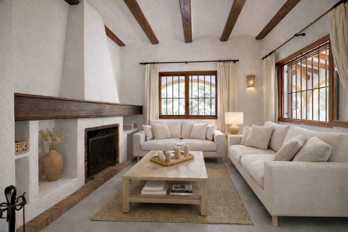 Salón con chimenea tradicional de estuco blanco y viga de madera oscura. Destacan las vigas vistas en el techo y los ventanales con marcos de madera y rejas. Salón con chimenea tradicional de estuco blanco y viga de madera oscura. Destacan las vigas vistas en el techo y los ventanales con marcos de madera y rejas.