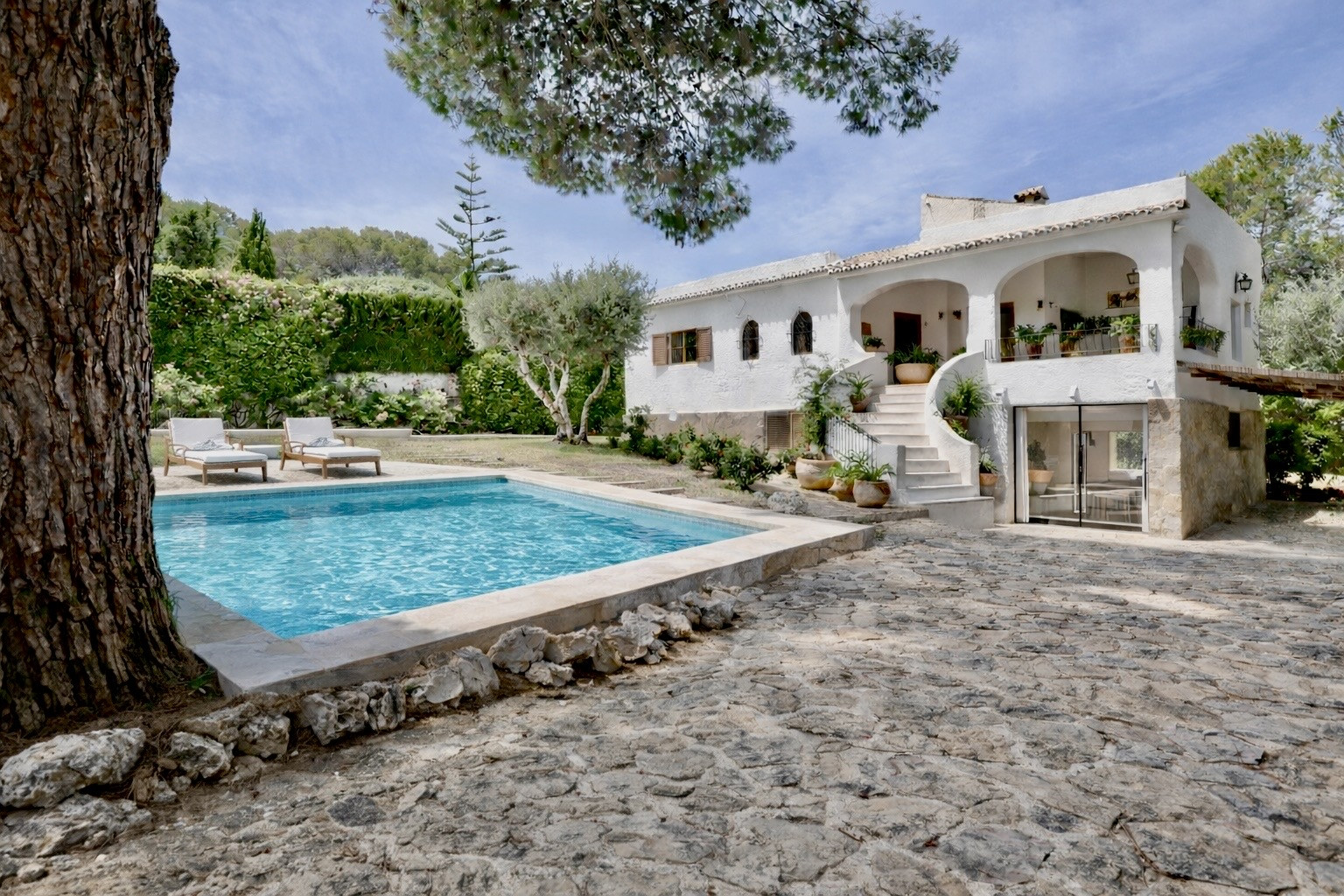 Vista exterior de una villa blanca tradicional con piscina privada, patio empedrado y escalera exterior. Rodeada de un exuberante jardín mediterráneo. Vista exterior de una villa blanca tradicional con piscina privada, patio empedrado y escalera exterior. Rodeada de un exuberante jardín mediterráneo.