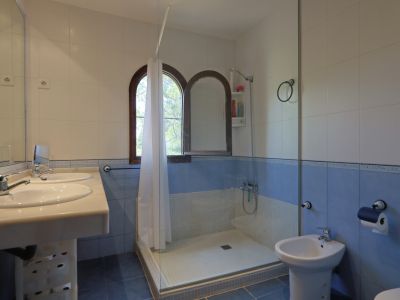 Baño con doble lavabo y encimera clara, paredes alicatadas en blanco y azul, y ducha con mampara de cristal. Destacan las dos ventanas arqueadas de madera que aportan luz natural.