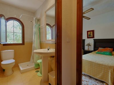 Baño con ducha, inodoro y lavabo de pedestal, destacando una ventana arqueada de madera con vistas a la vegetación. Se conecta visualmente con un dormitorio adyacente.