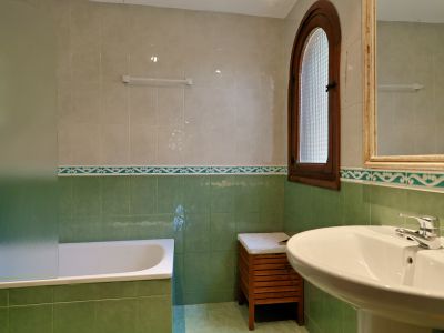 Baño con paredes alicatadas en tonos verde y beige, bañera con mampara de cristal esmerilado y lavabo de pedestal. Ventana de madera arqueada con cristal texturizado.