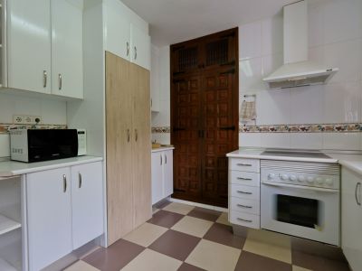 Cocina equipada con armarios blancos, microondas y horno integrado. Destaca la puerta de madera oscura con paneles y el suelo de baldosas ajedrezadas.