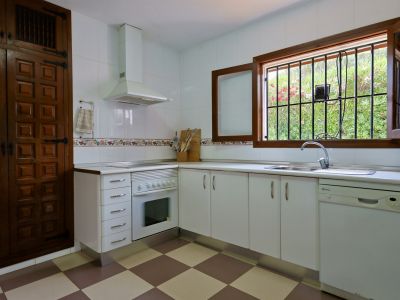 Cocina funcional con armarios blancos, suelo de baldosas a cuadros y una ventana de madera con rejas que ofrece vistas al jardín. Incluye fregadero de acero y lavavajillas.