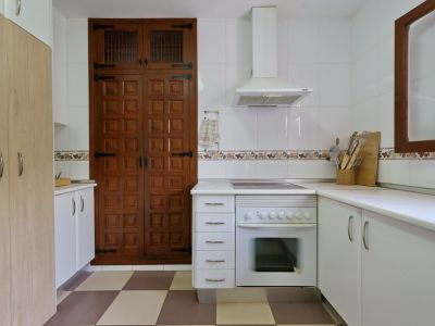 Cocina funcional con horno integrado, campana extractora y azulejos blancos. Destaca un gran armario de madera con puertas paneladas y herrajes tradicionales, sobre un suelo ajedrezado.