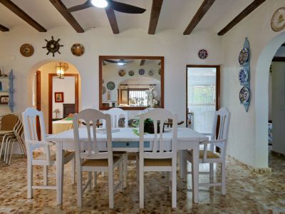 Comedor con mesa y sillas blancas de madera, suelo de baldosas con patrón y techo con vigas de madera oscura. Un espejo enmarcado refleja la estancia y su decoración.