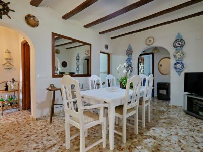 Comedor con mesa y sillas de madera blanca, suelo de terrazo y techo con vigas vistas. Arcos decorativos y espejos amplían el espacio.