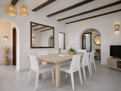 Comedor luminoso con mesa de madera clara para ocho personas y sillas blancas. El techo presenta vigas de madera oscura y lámparas colgantes de mimbre, junto a un gran espejo con marco.