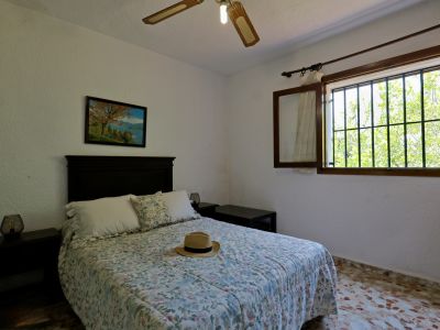 Dormitorio acogedor con cama doble, ropa de cama floral y cabecero de madera oscura. Ventana enrejada con vistas a la vegetación exterior y ventilador de techo.