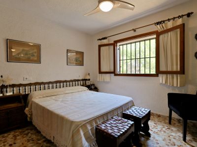 Dormitorio con cama doble, cabecero de madera y suelo de baldosas. La ventana con contraventanas y rejas ofrece luz natural y vista a la vegetación exterior.