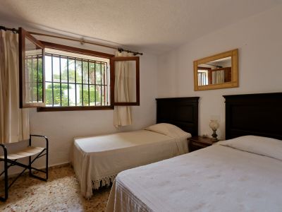 Dormitorio con dos camas individuales de cabeceros de madera oscura, mesita de noche compartida y ventana con rejas hacia el jardín. Suelo de terrazo y espejo con marco trenzado.